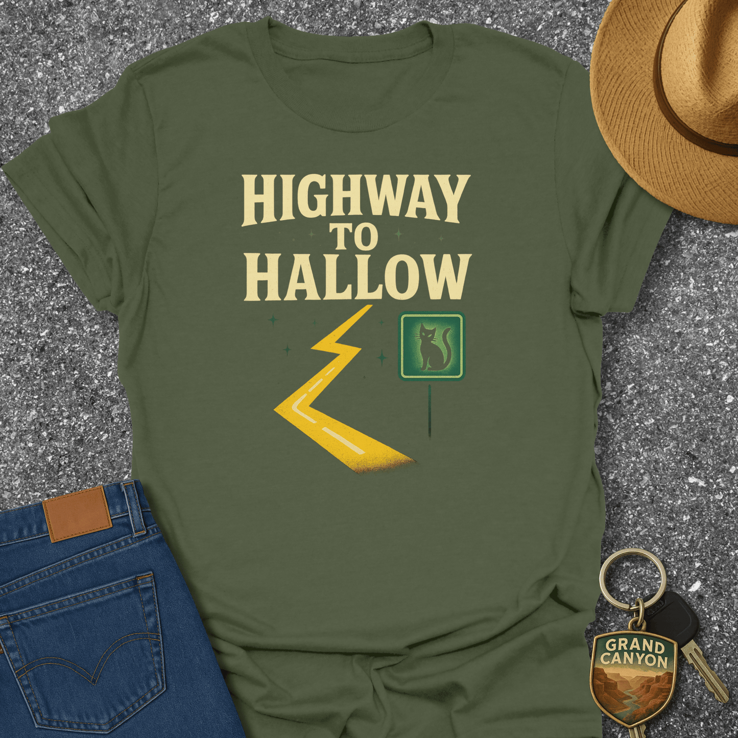 Green t-shirt with 'Highway to Hallow' design, placed on a textured surface with jeans, a hat, and keychain.