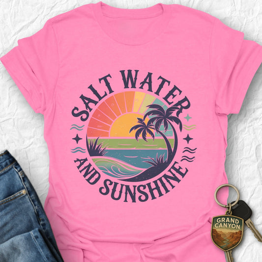 Pink t-shirt with 'Salt Water and Sunshine' design on a textured surface with jeans and a keychain.