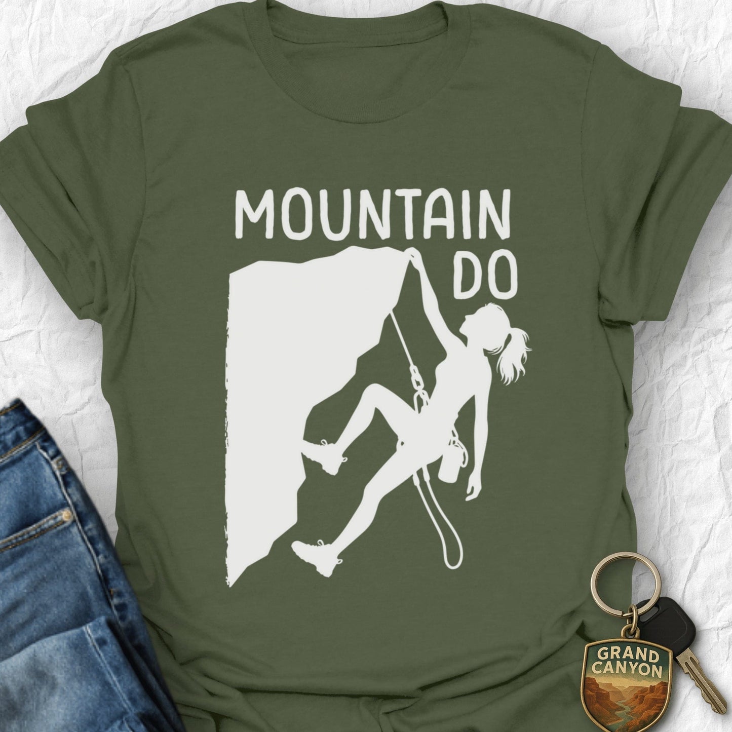 Green t-shirt with white graphic of a climber and 'Mountain Do' text on a textured surface.