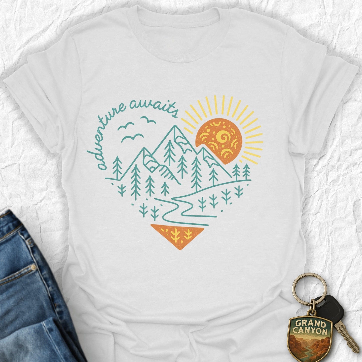 White t-shirt with mountain and sun design, next to jeans and a keychain on a white background
