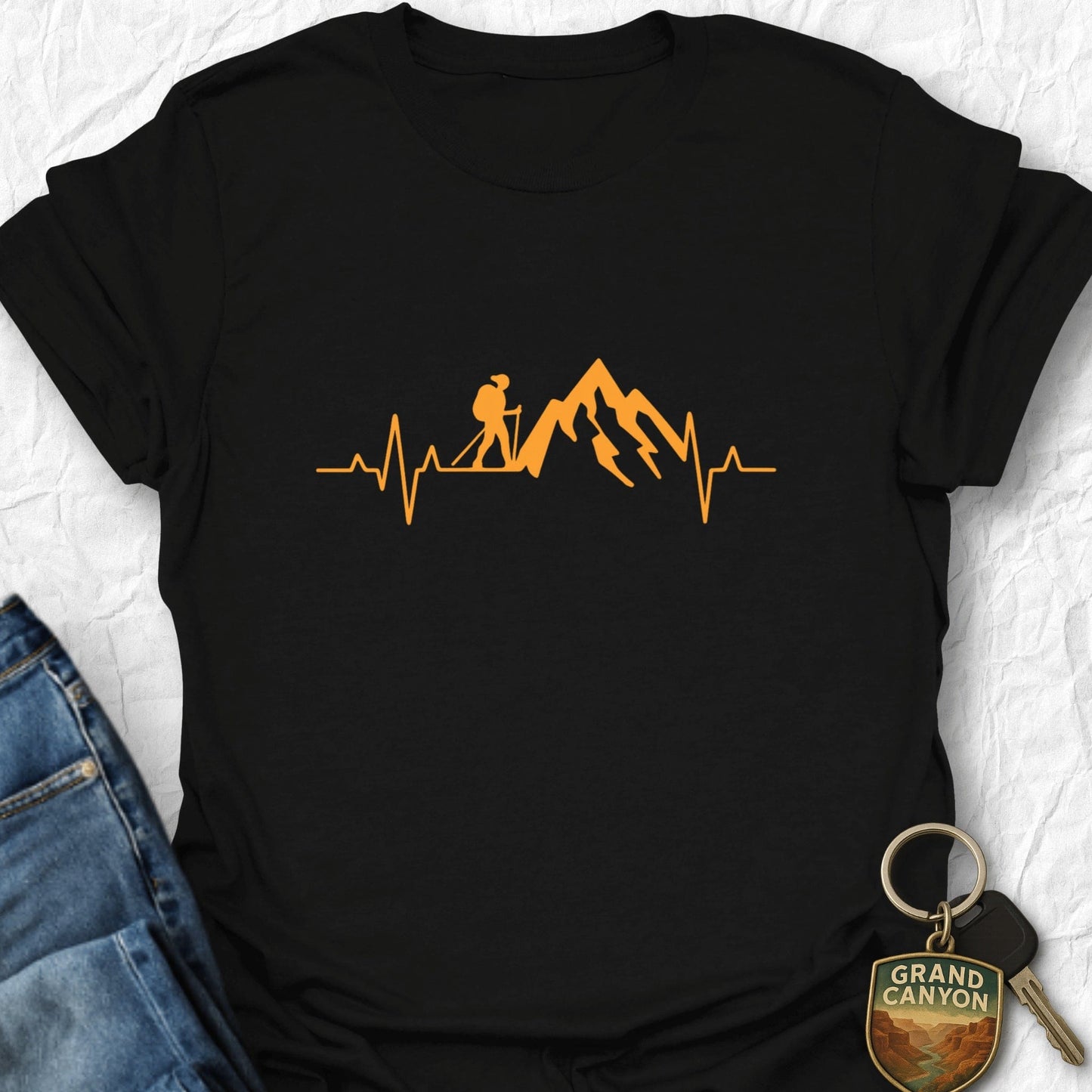 Black t-shirt with orange mountain and heartbeat design, next to jeans and a keychain on a white background